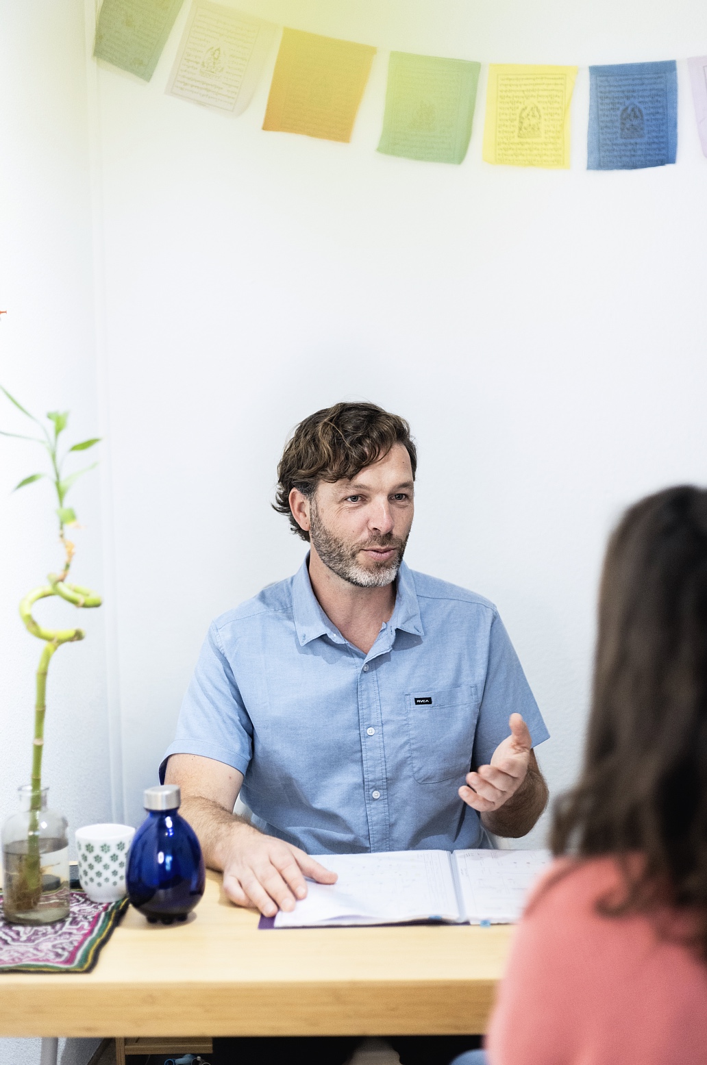 Man in a light blue shirt sits at a wooden desk, gesturing as he speaks to someone off-camera with papers open in front of him. A blue bottle and plant are on the desk beneath colorful wall flags.