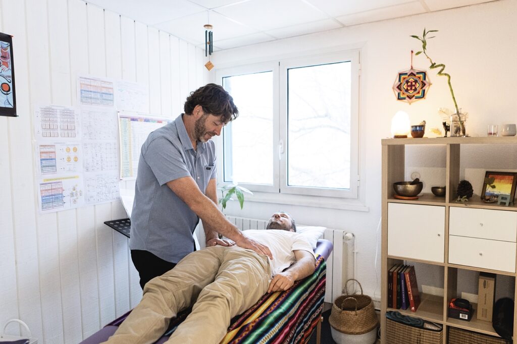 Therapist performing a back adjustment on a patient lying on a treatment table in a bright, tidy clinic with charts on the wall and a striped blanket on the table.