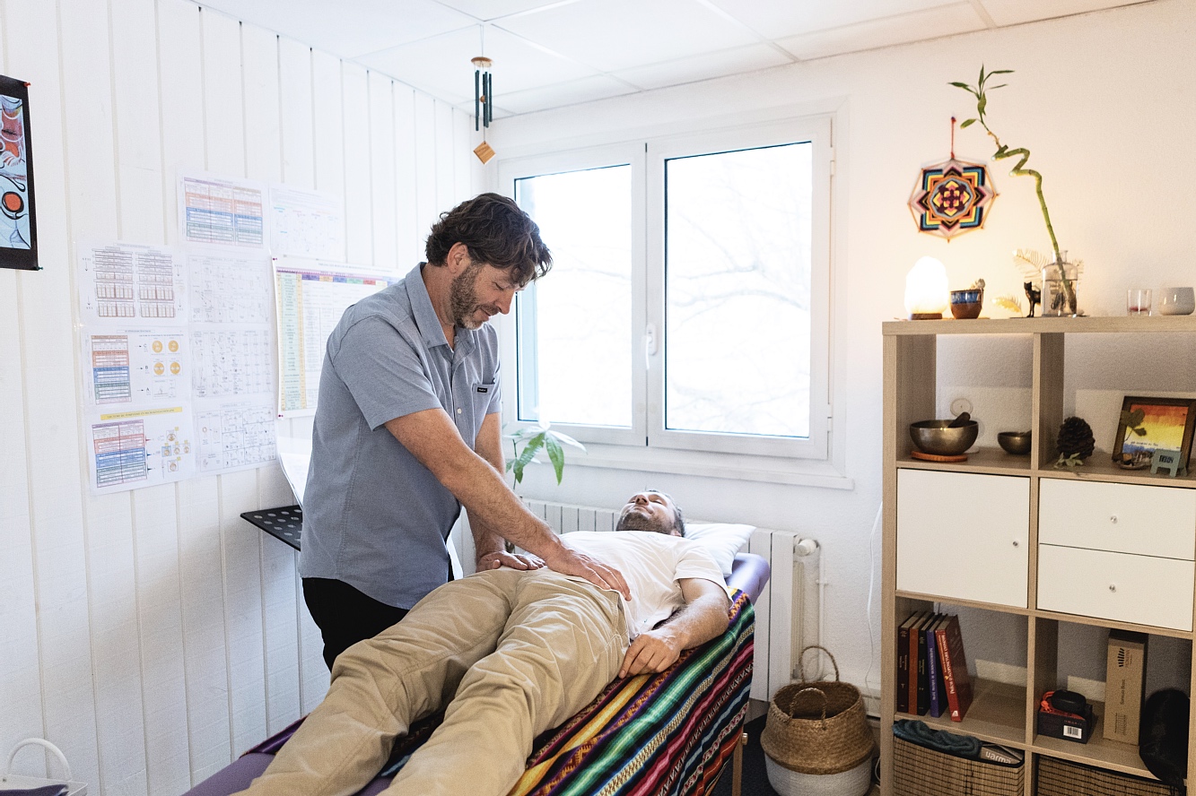 Therapist performing a back adjustment on a patient lying on a treatment table in a bright, tidy clinic with charts on the wall and a striped blanket on the table.