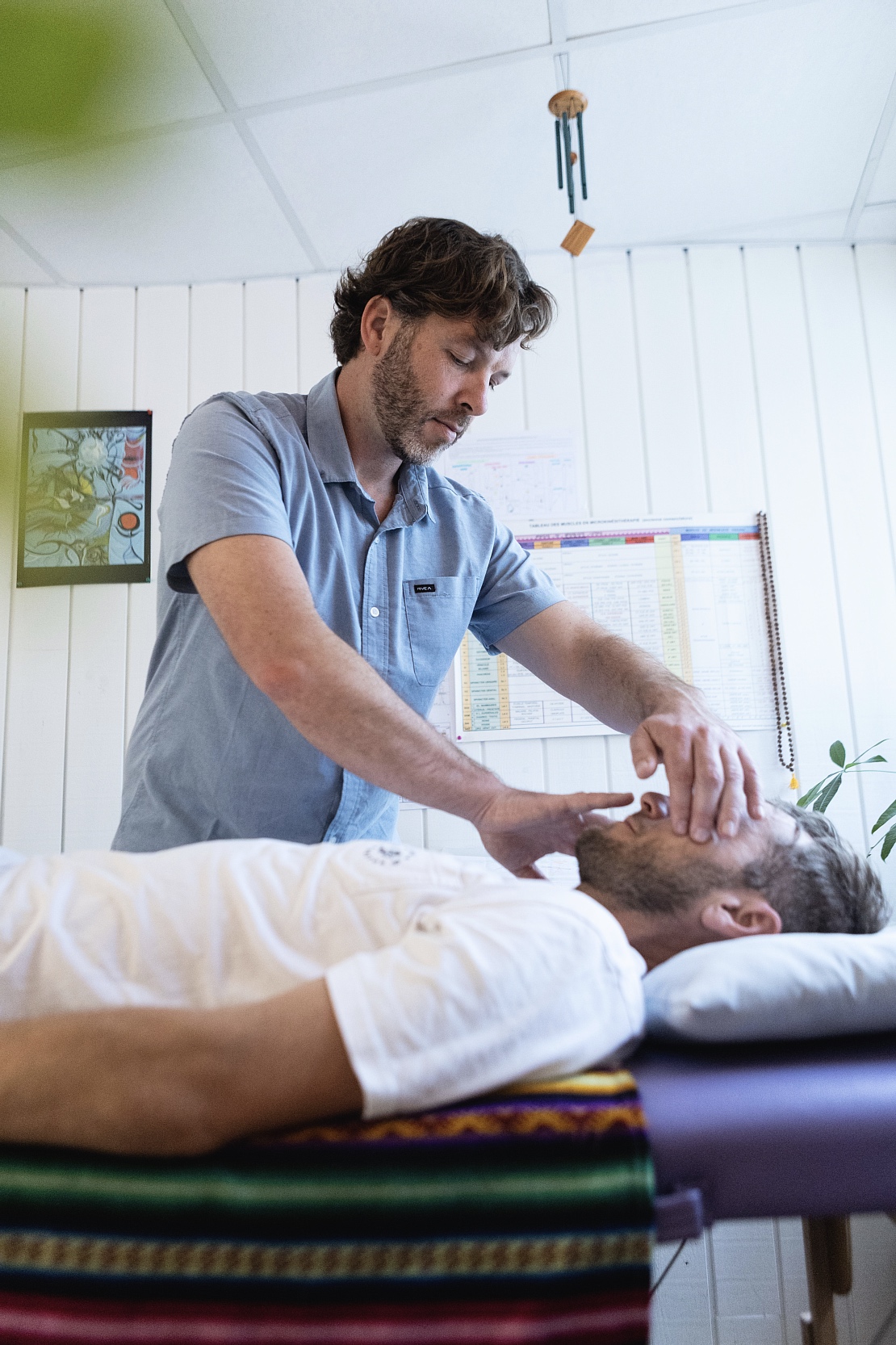 Male therapist stands beside a patient lying on a padded table, using his hands to perform a facial/jaw treatment in a clinical room.