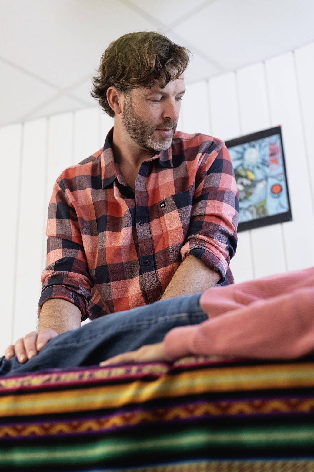 Man in a red plaid shirt leaning over a stack of colorful blankets on a table indoors.