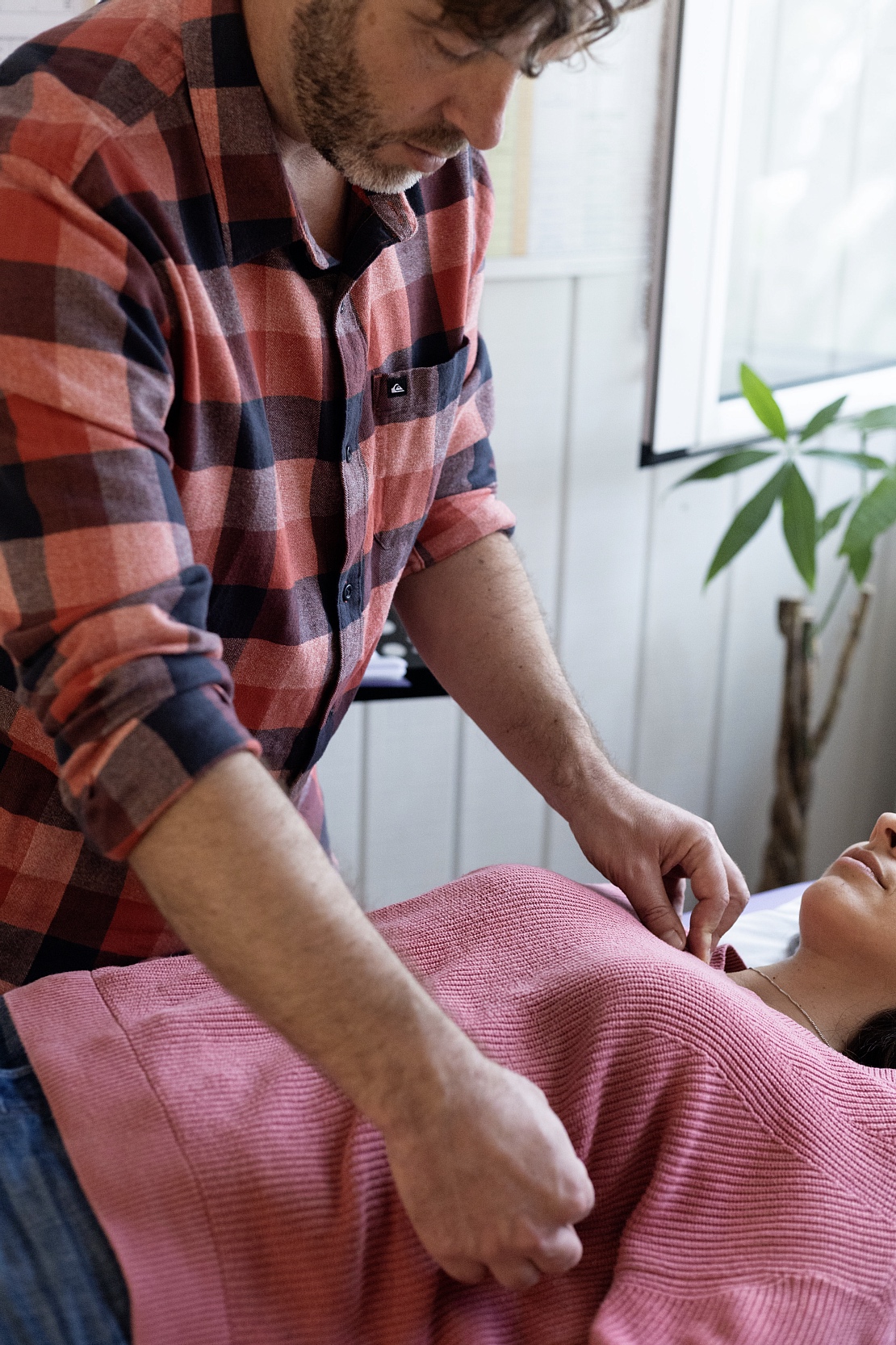 Caregiver in a red plaid shirt leaning over a patient, adjusting a pink blanket on a bed.