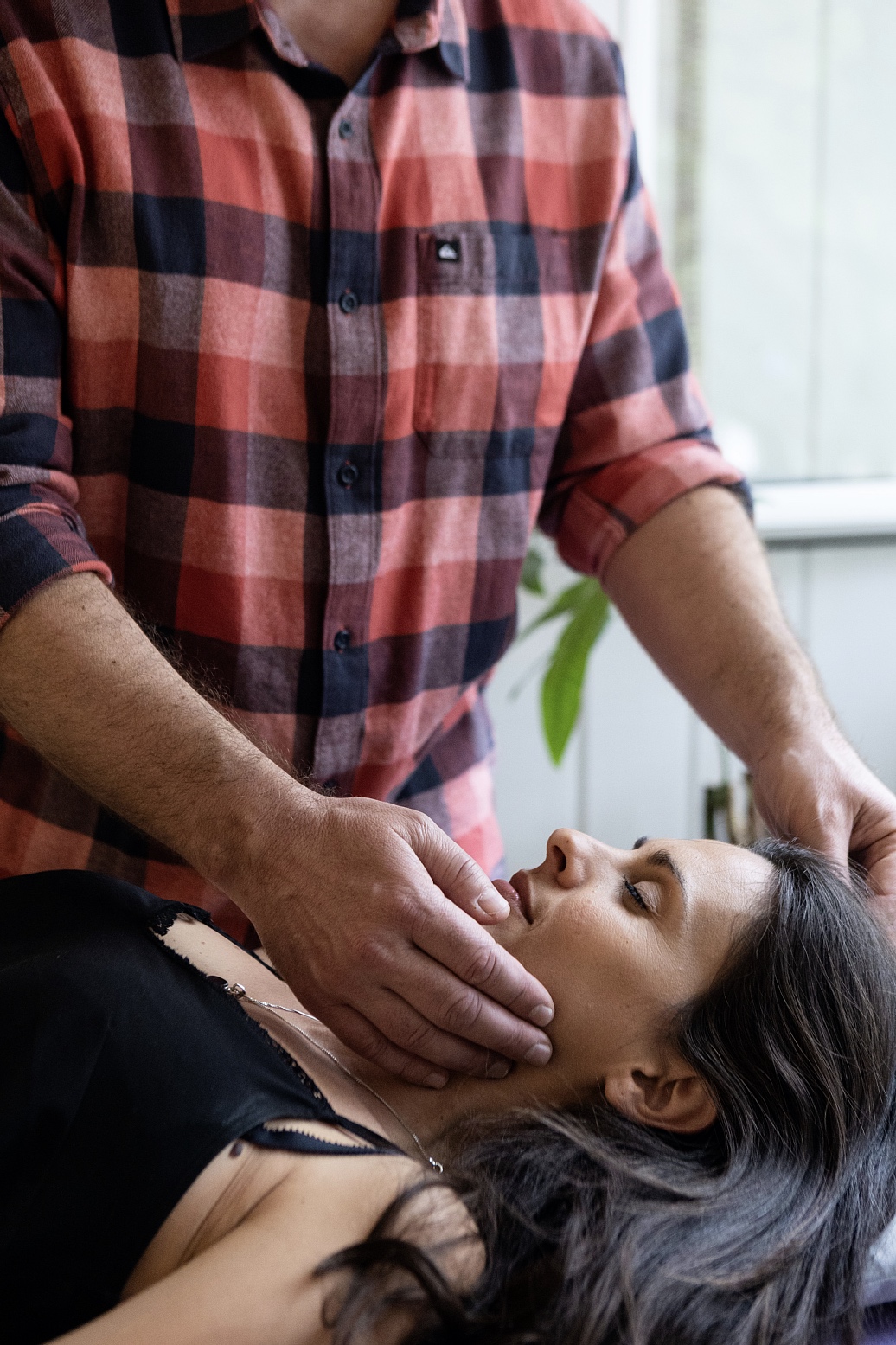 Man in a red plaid shirt supports a woman's head and neck as she lies on her back.