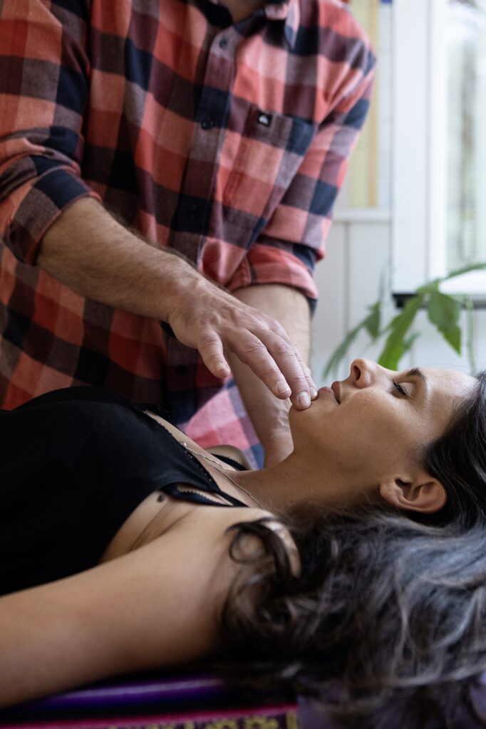 Man in a red plaid shirt leans over a woman lying down, his fingers near her lips during a soothing treatment.