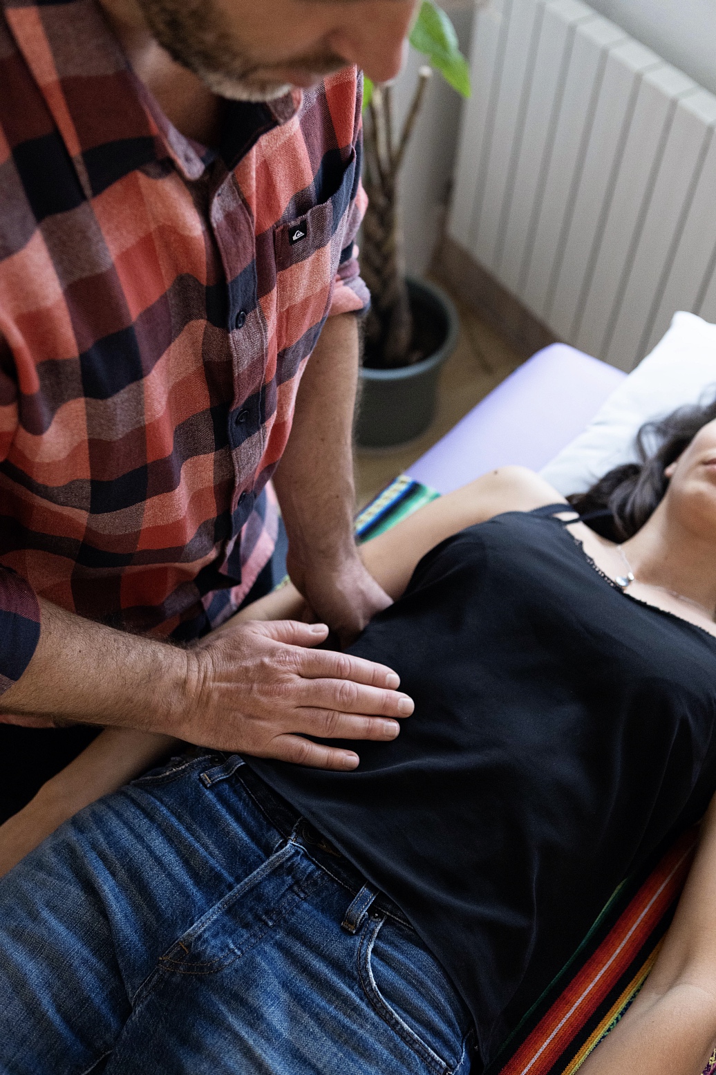 Man in a red plaid shirt performing chest compressions on a woman who is lying on a bed or examination table, illustrating CPR.